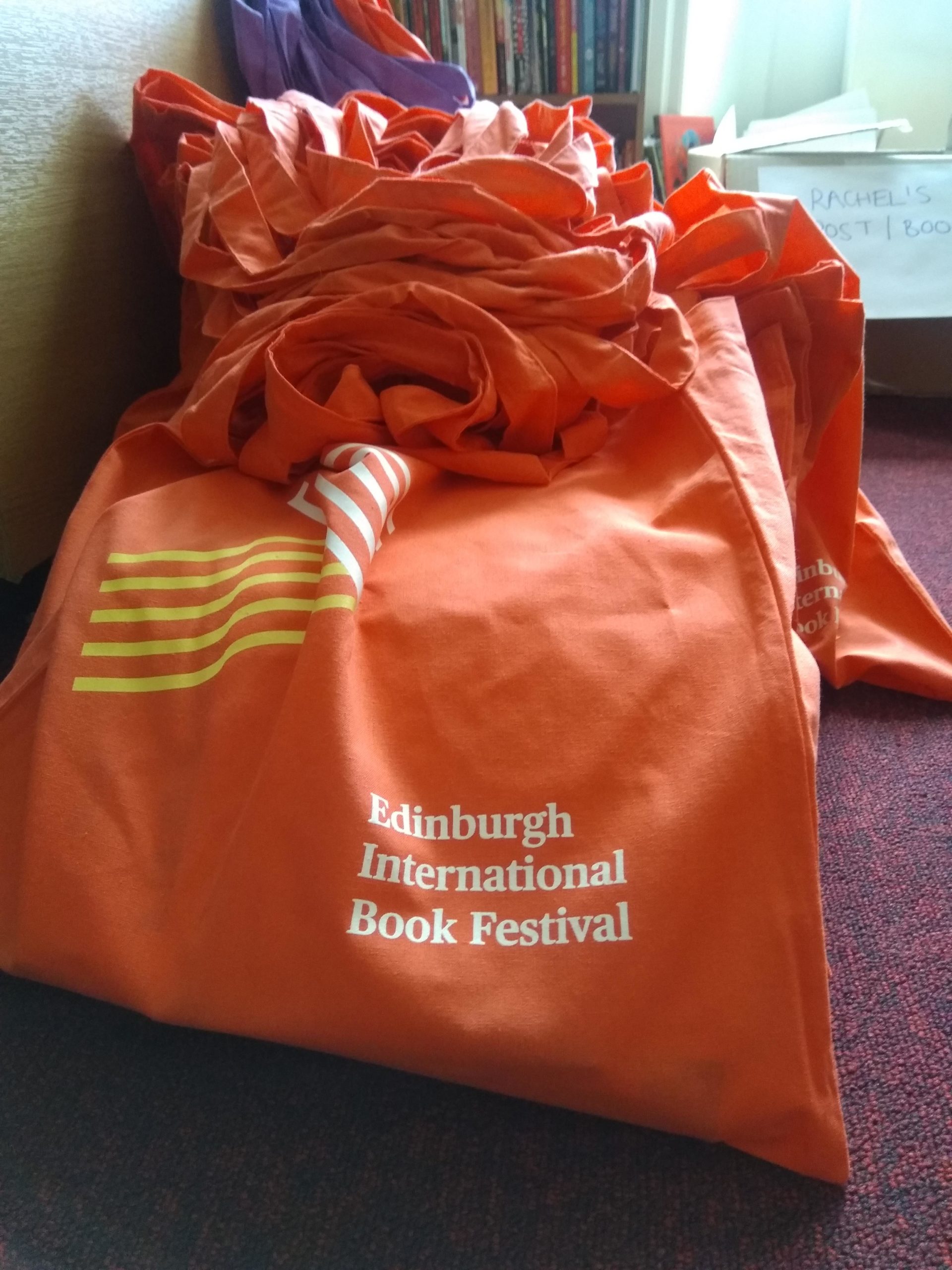 A stock of Edinburgh International Book Festival tote bags stacked against a bookshelf