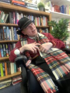 Mark McManus wearing his trademark black hat, sitting in his electric wheelchair in front of a bookcase.